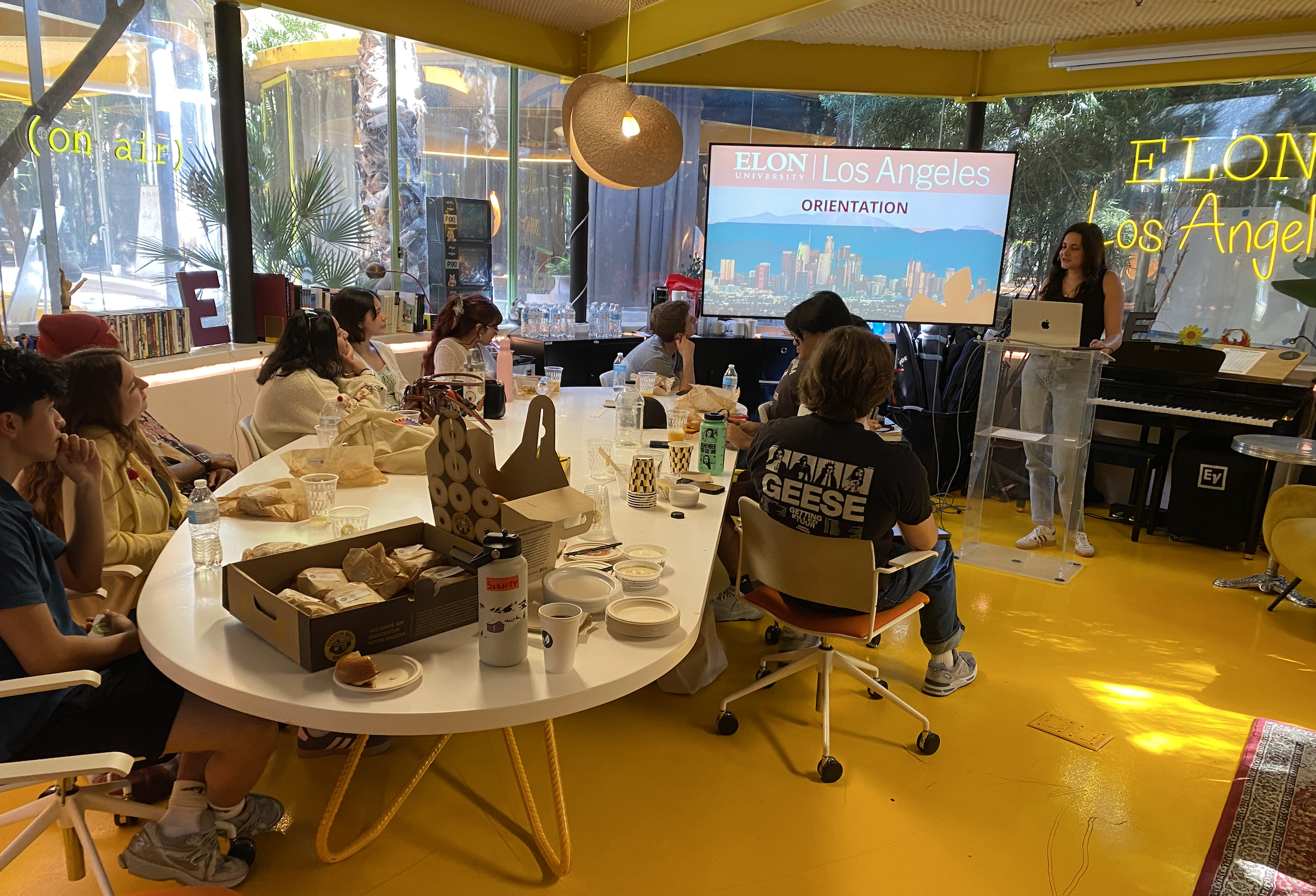 Students sit around tables listening to a presentation during an Elon University Los Angeles orientation session in a bright, modern room. A presenter stands at the front beside a screen displaying the orientation slide, while food and drinks are set out on the tables.