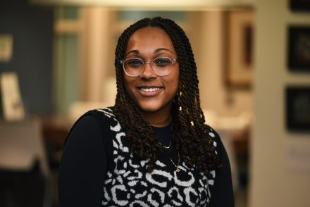 A woman with glasses and dark, long locs smiles at the camera. She is wearing a black long-sleeve shirt with a patterned white circle design over.
