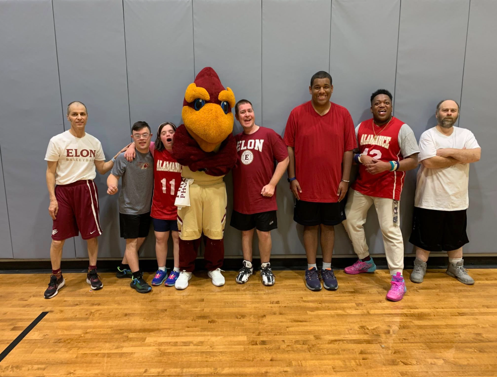 Seven men and a large bird mascot wearing Elon apparel stand together on a gym floor in front of a gray padded wall, smiling and posing with their arms around each other. Several wear red Elon shirts or jerseys, creating a spirited team photo atmosphere.