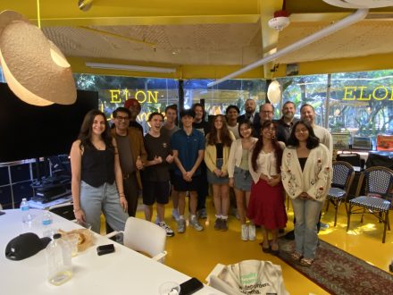 A group of students and staff stand together smiling in a bright room during an Elon University Los Angeles orientation event. Tables, chairs, and windows with “ELON” signage are visible, creating a welcoming group setting.