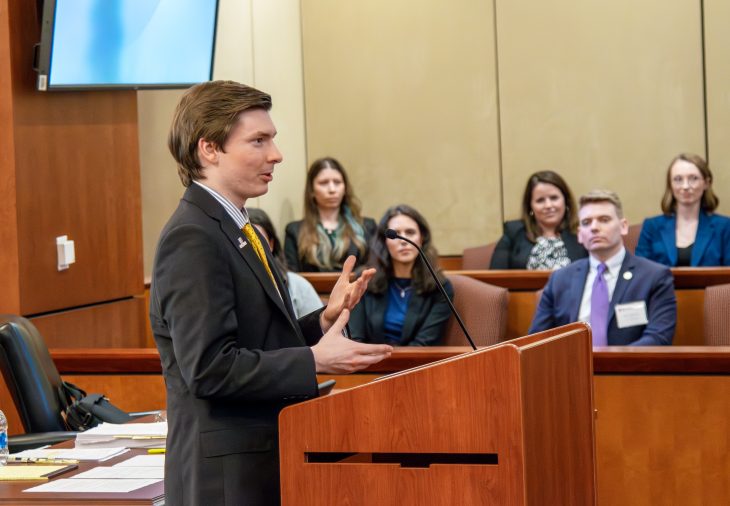 a man speaks from behind a podium in a crowded courtroom. He is wearing a suit and gesturing with his hands.