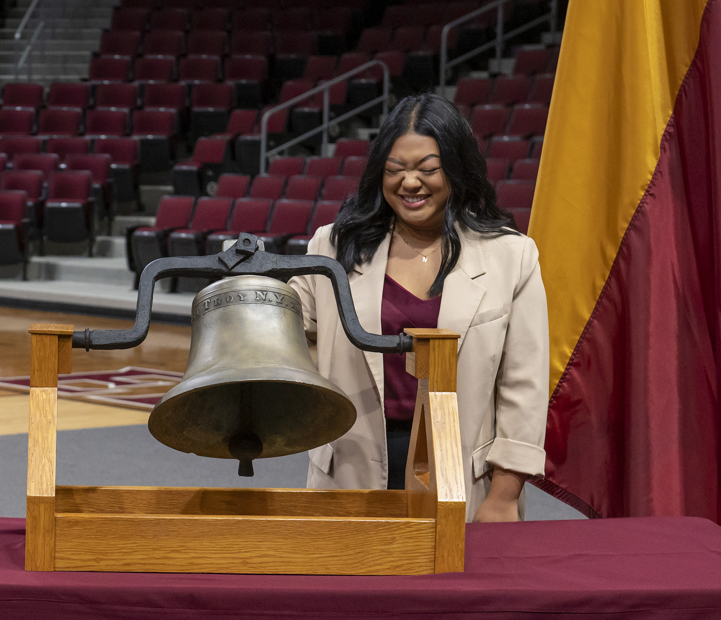 Student rings the Elon University bell during a campus ceremony in an arena with maroon seating.