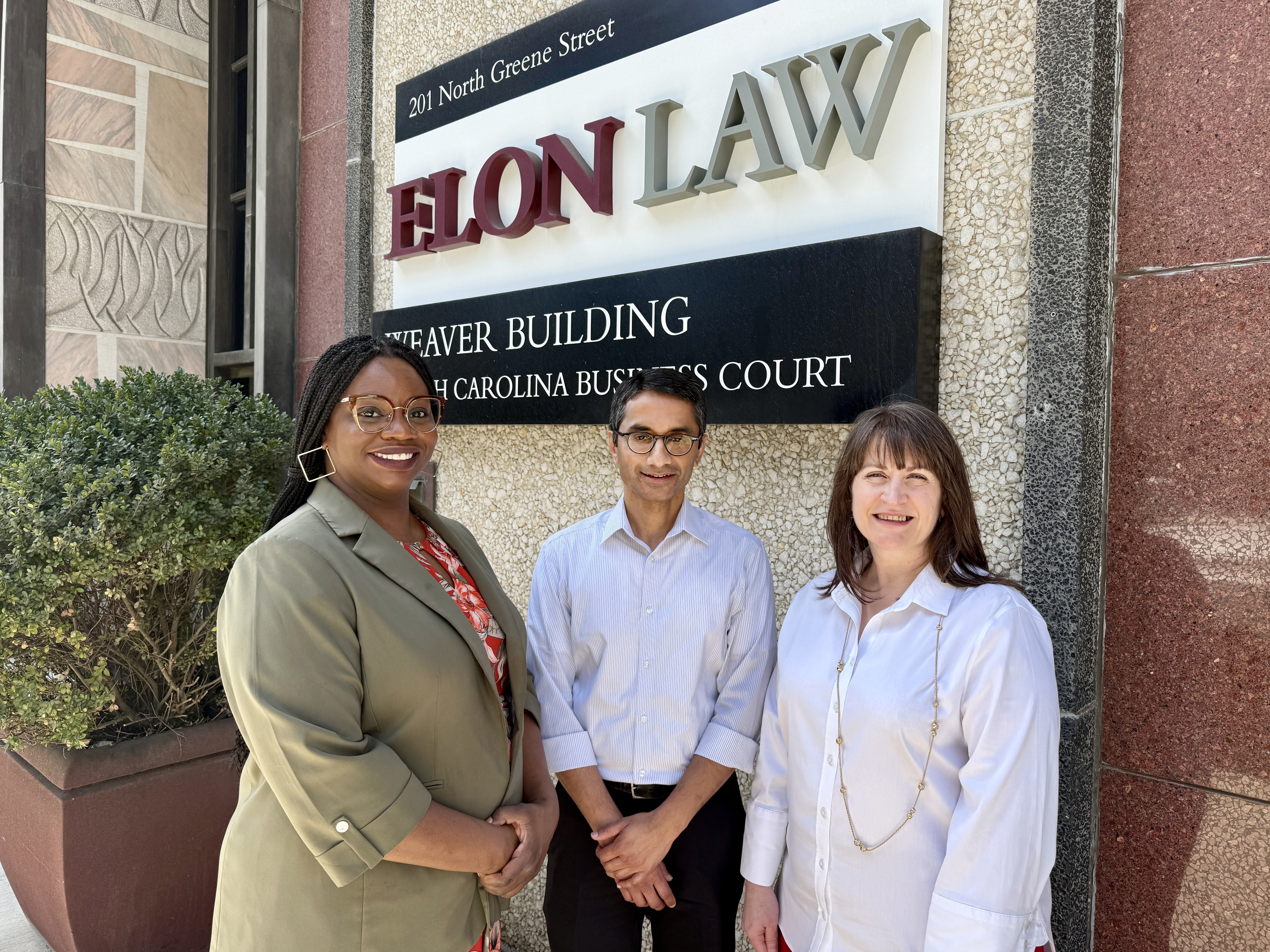 Two women and a man in front of the Elon Law exterior sign on 201 N. Greene St. in downtown Greensboro