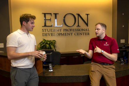 Two people talk at the front desk of the Elon Student Professional Development Center, one holding a coffee while they chat.