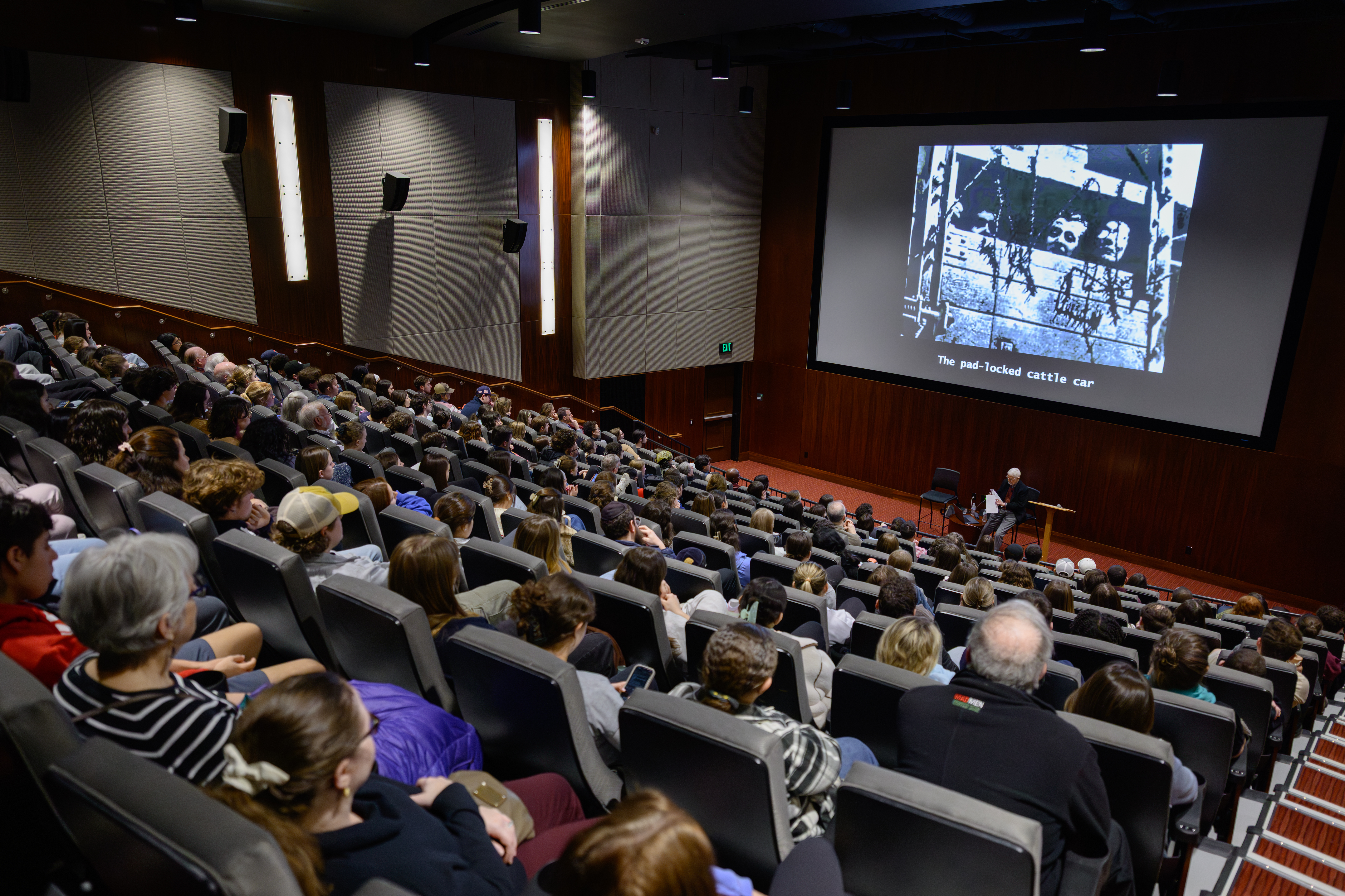 A large audience sits in a tiered lecture hall watching a presentation with a projected historical image and the caption “The pad-locked cattle car.” A speaker sits at the front of the room beneath the screen, suggesting a formal educational or remembrance event.
