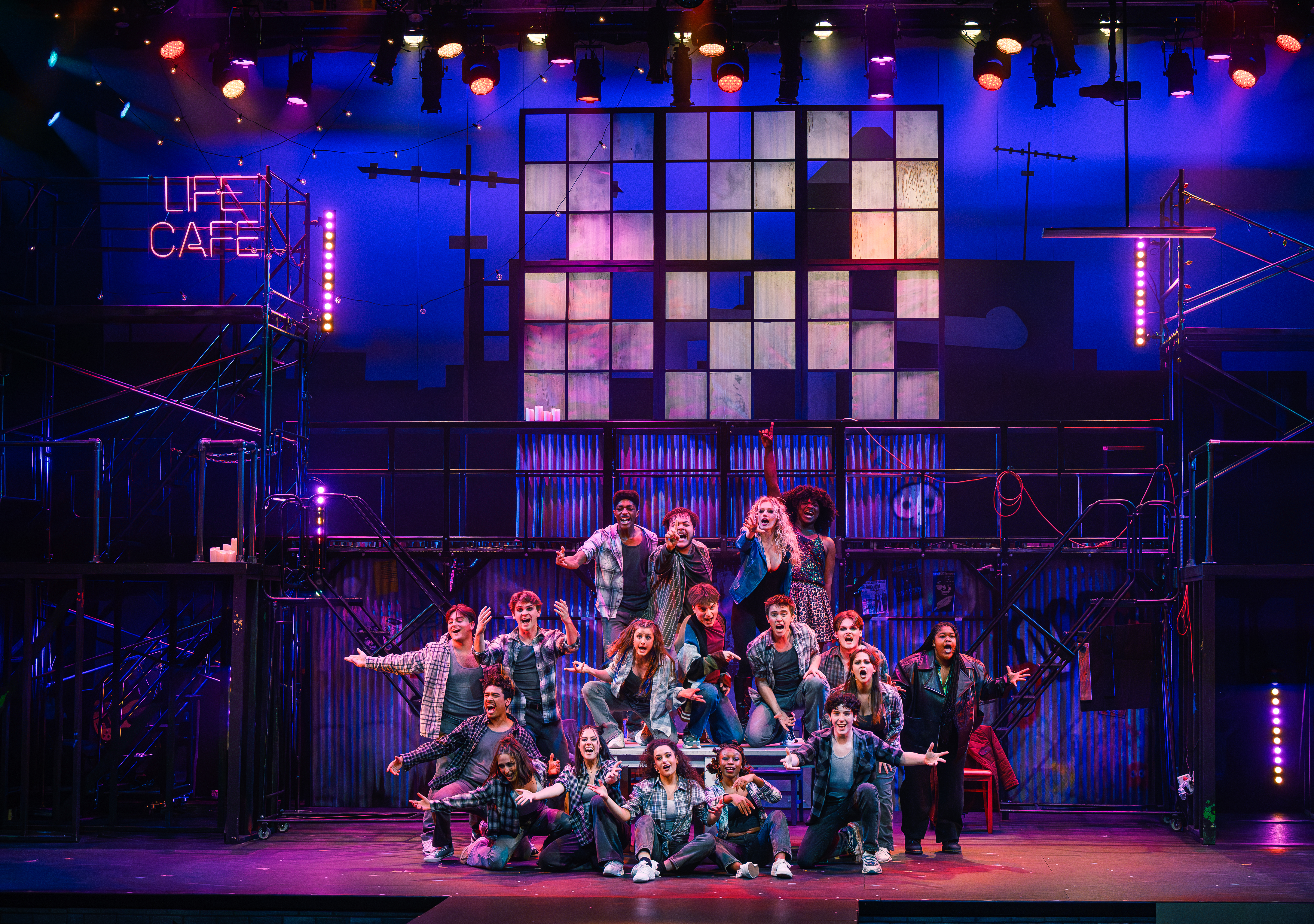 A group of performers on stage in front of a set featuring scaffolding and a checkerboard pattern of lights. A sign reads "Life Cafe." The lighting is vibrant, with blue and purple hues. The scene evokes a lively musical theater performance.