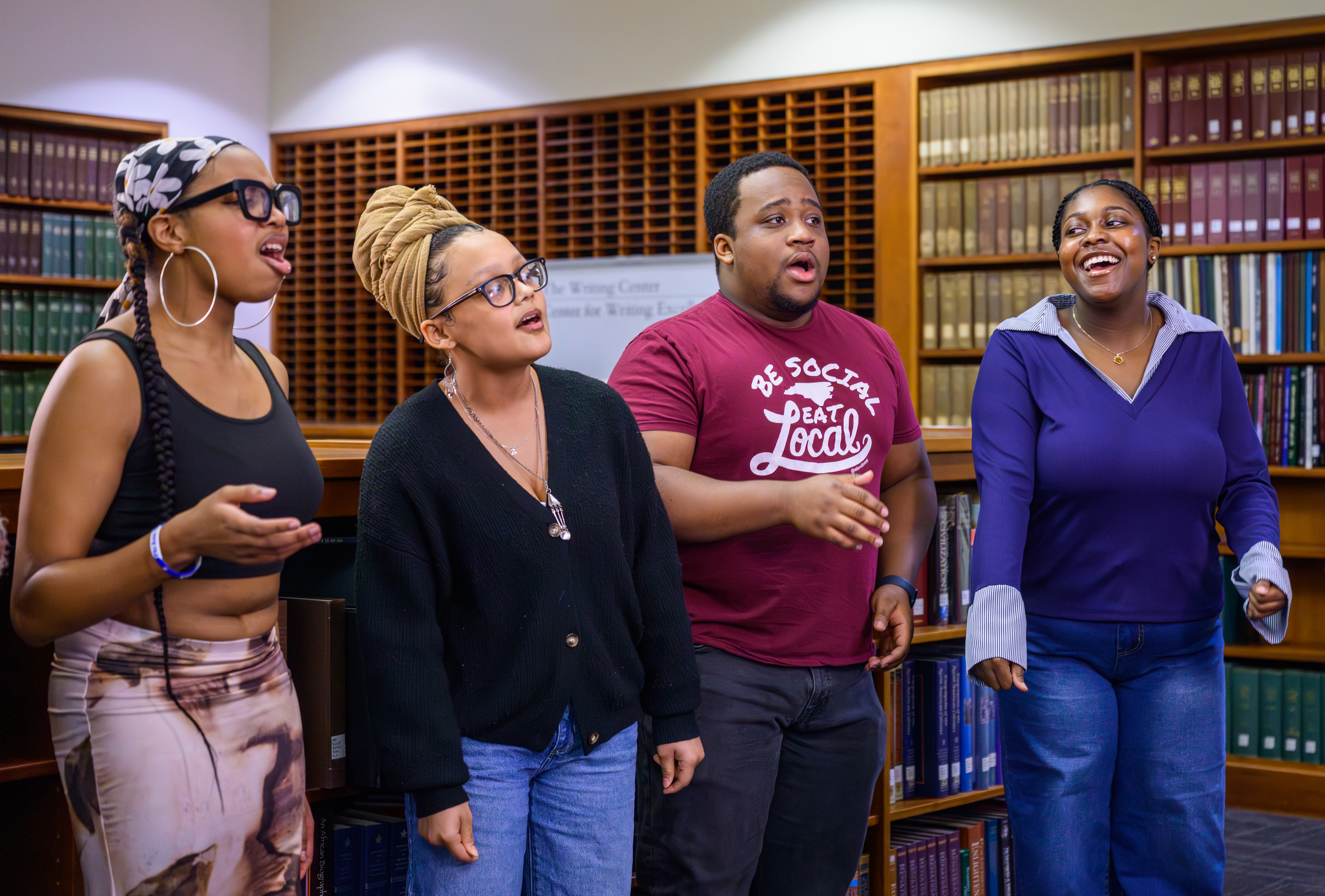 A group of four people are singing enthusiastically in a library or similar setting. They are standing in front of shelves filled with books. All are casually dressed and appear engaged in the activity.