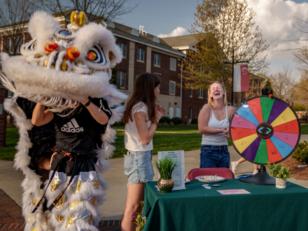 White lion dance costume interacts with students at an outdoor campus event, as two students laugh beside a table with a colorful prize wheel.