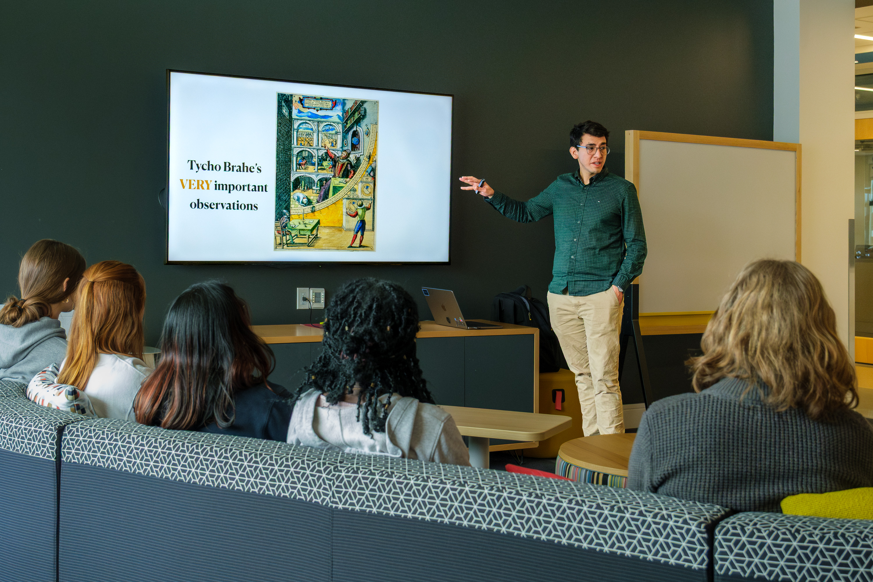A student stands at the front of a lounge-style classroom presenting slides on a screen while several students sit on couches and watch. The slide reads “Tycho Brahe’s VERY important observations,” suggesting a presentation related to astronomy or science.