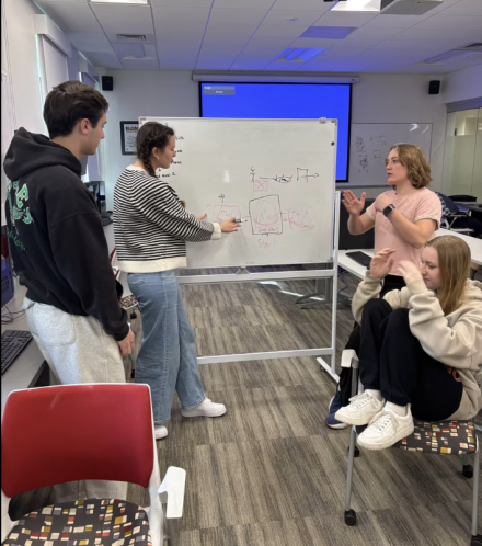 Group of four students writing and brainstorming around a white board. 