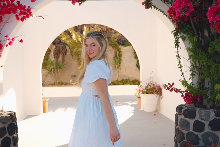 A woman in a light blue dress stands beneath a white archway surrounded by bright pink flowers, smiling toward the camera. Sunlight illuminates the scene, creating a warm, picturesque outdoor setting.