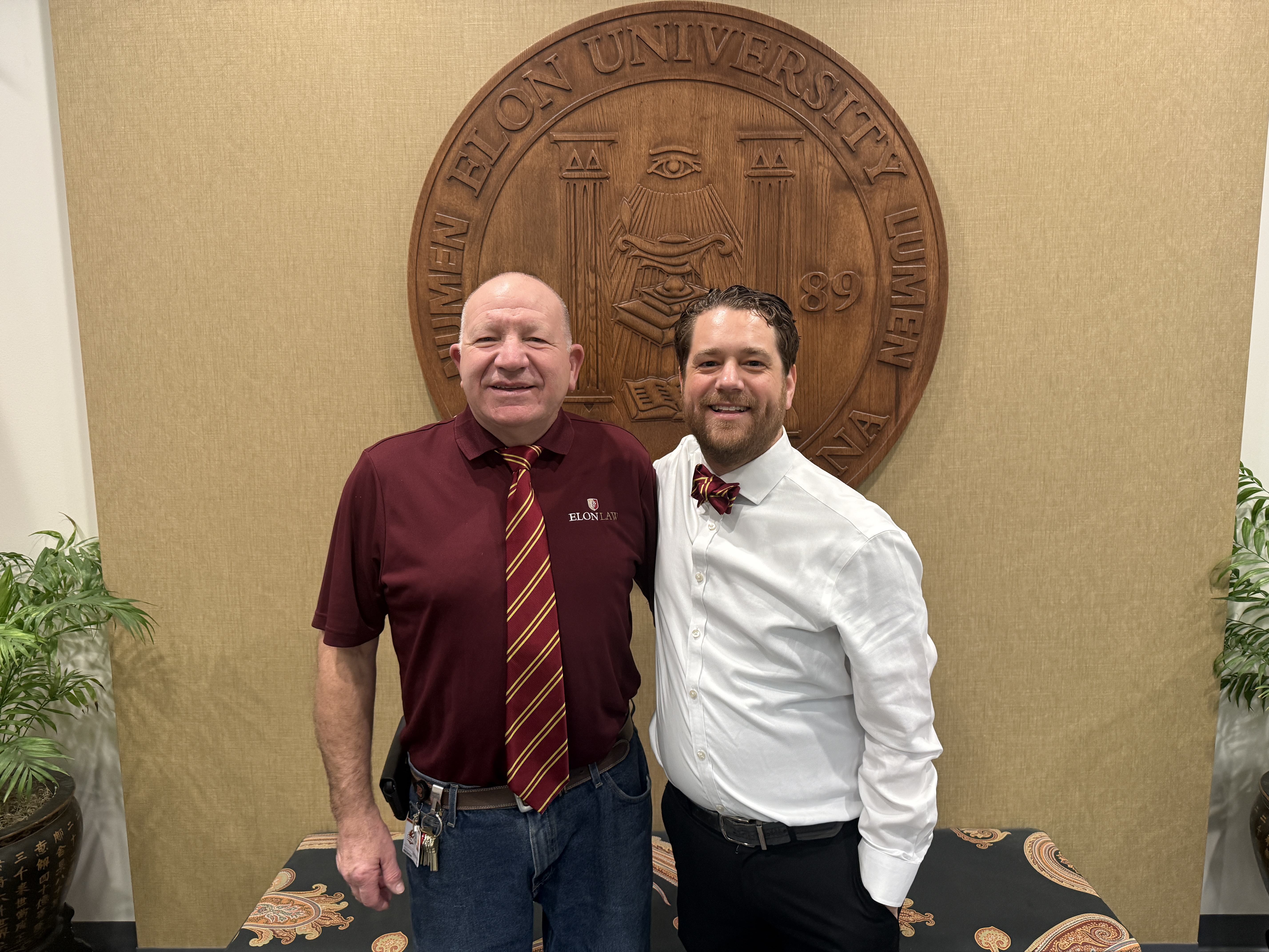 Two men smiling, wearing maroon and gold ties, in front of the Elon University seal in the law school dean's suite