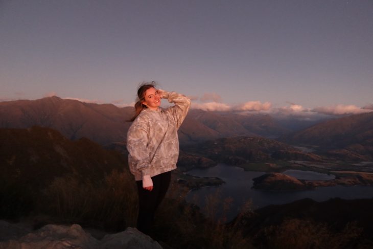 A woman stands on a mountain overlook at sunset, smiling with a lake and rolling hills stretching out below. The warm evening light casts a soft glow over the landscape and her face.