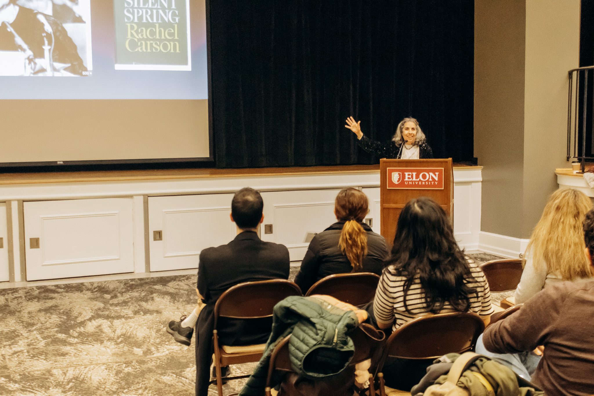Women at podium delivering speech with people sitting in rows of chairs in front of her.