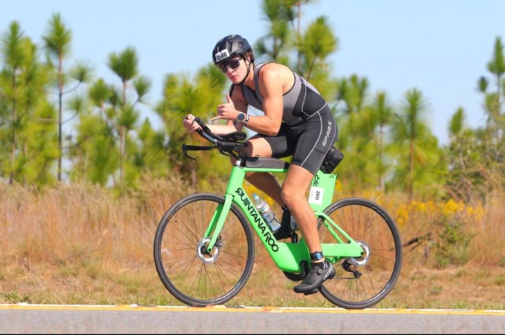An athlete rides a road bike in a triathlon, wearing a helmet and aerodynamic gear while leaning forward in a racing position. The scene shows a rural roadside with trees in the background.