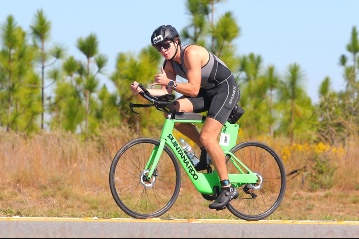 An athlete rides a road bike in a triathlon, wearing a helmet and aerodynamic gear while leaning forward in a racing position. The scene shows a rural roadside with trees in the background.