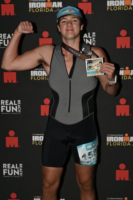 A young athlete poses in front of an Ironman Florida backdrop, flexing his arm and holding a finisher medal. He wears a triathlon suit and race bib, celebrating his completion of the event.