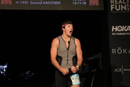 A young athlete crosses the finish line at an Ironman event, wearing a triathlon suit and race bib, celebrating with an excited expression.