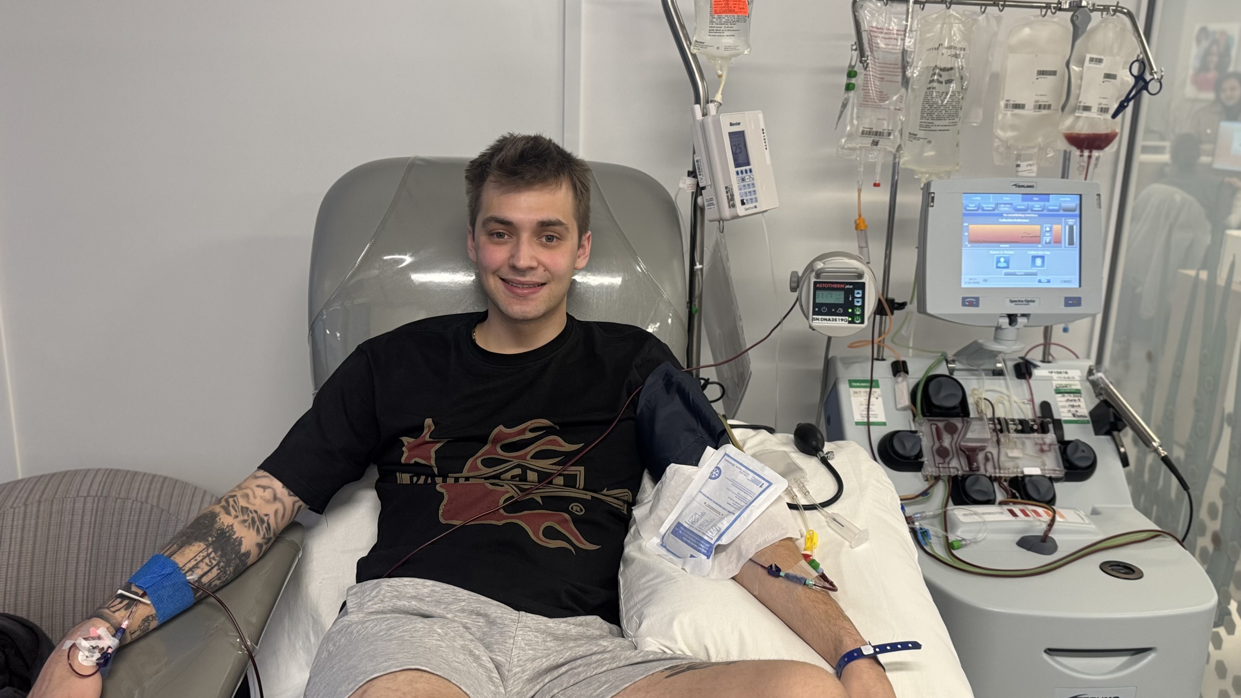 A young man sits in a medical chair smiling while connected to blood donation equipment, with tubes running from his arm to a nearby machine. Medical monitors and collection bags are visible beside him, indicating he is donating blood or platelets.