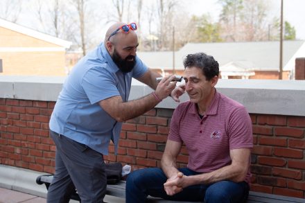 Justin Clar wearing a blue polo shaving off Joel Karty's eyebrows with an electric razor