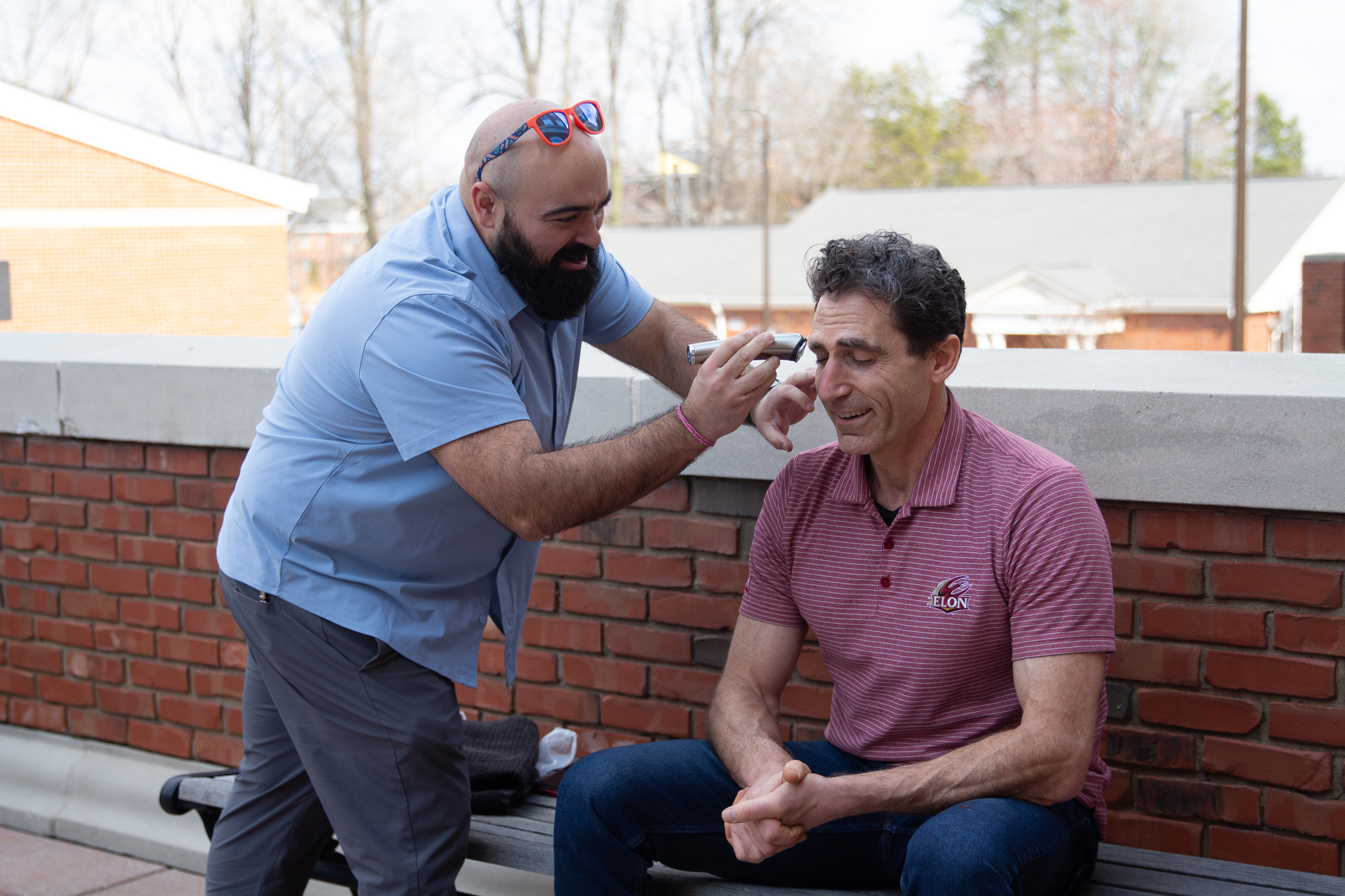 Justin Clar wearing a blue polo shaving off Joel Karty's eyebrows with an electric razor