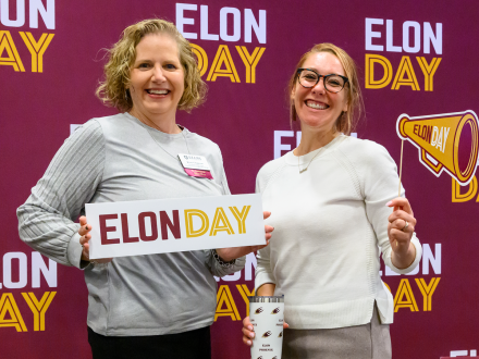 Two women smile in front of an Elon Day backdrop, holding an “Elon Day” sign and megaphone prop.