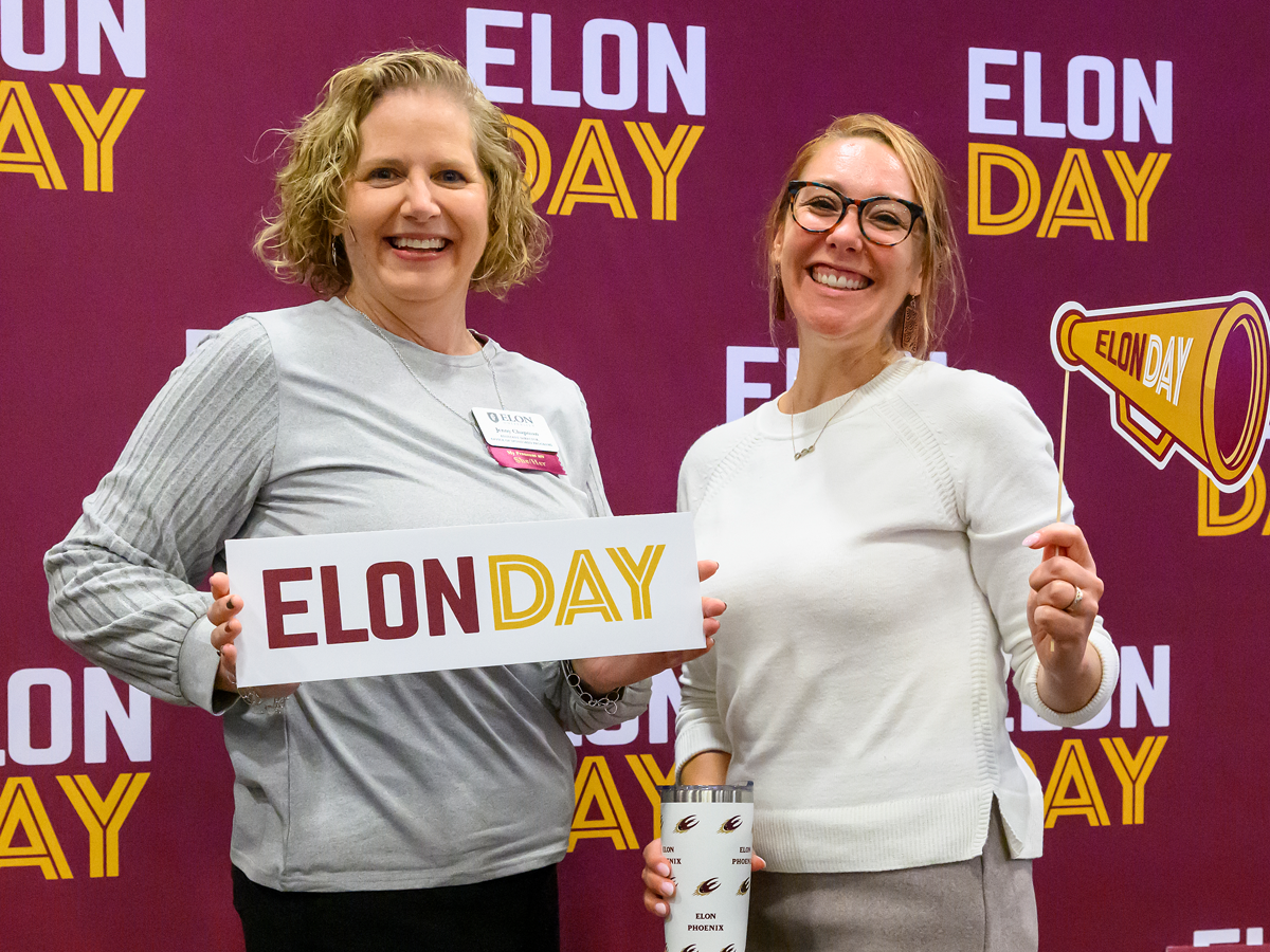 Two women smile in front of an Elon Day backdrop, holding an “Elon Day” sign and megaphone prop.