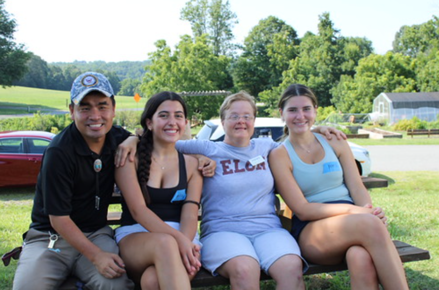 Four people sit closely together on a bench outdoors, smiling with their arms around each other. Trees, cars, and a grassy area appear in the background, suggesting a relaxed campus or park setting.
