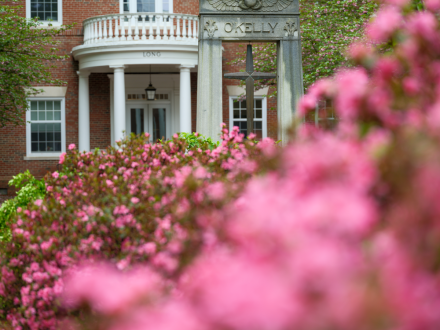 O’Kelly monument and cross in front of Long Building, framed by blooming pink flowers.