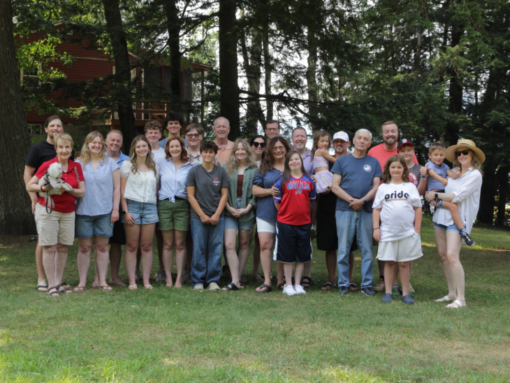A large group of people of various ages stand together on a grassy lawn in front of trees and a cabin, posing for a group photo. The scene suggests a family gathering or reunion in a wooded outdoor setting.