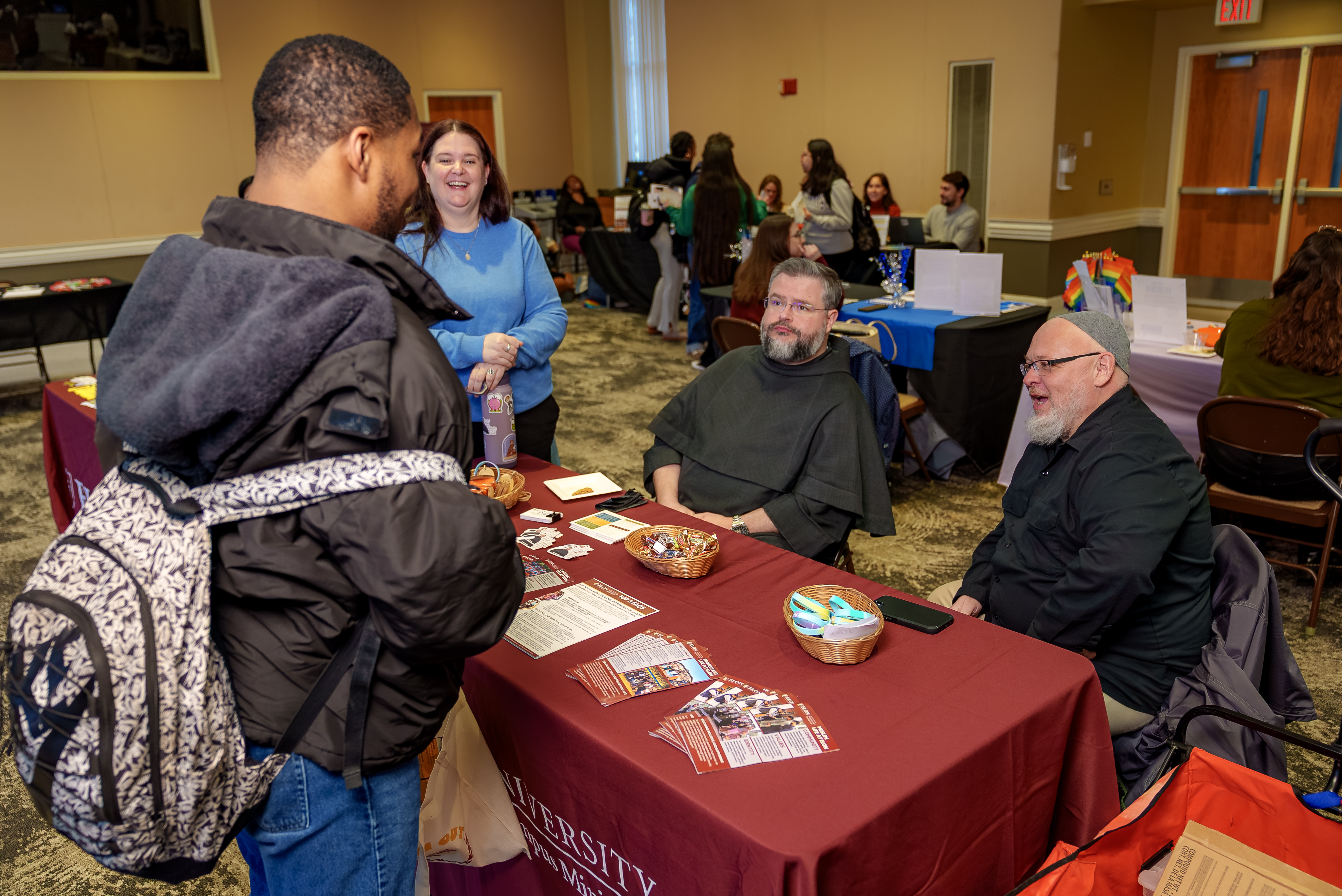 A student with a backpack speaks with two seated representatives at a table covered with informational materials during a campus event. Other attendees and tables are visible in the background, suggesting a fair or networking session.