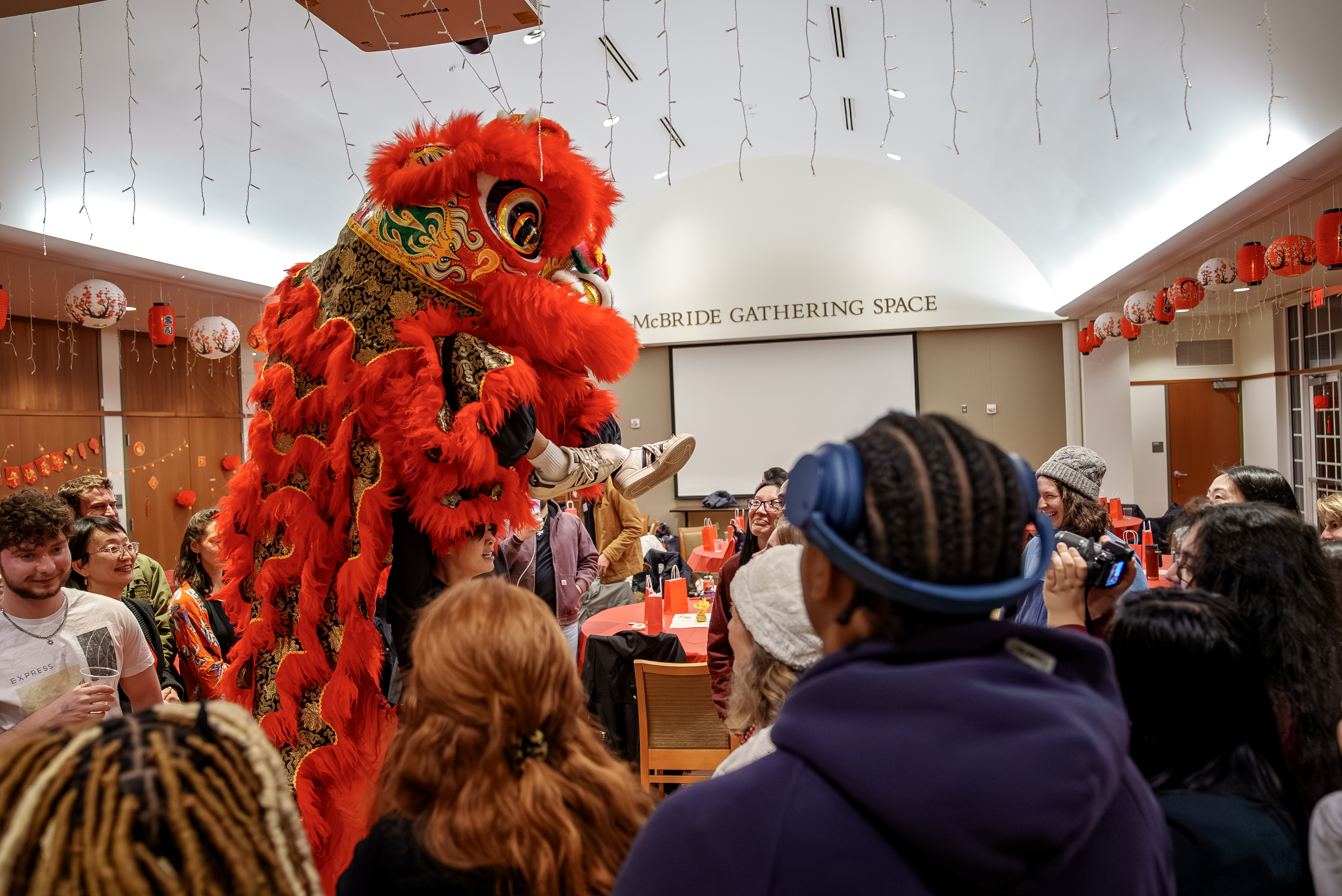 A vibrant red and gold lion costume is being worn by performers in a crowded room, engaging with an audience. The ceiling is decorated with string lights and red lanterns. A sign reads 