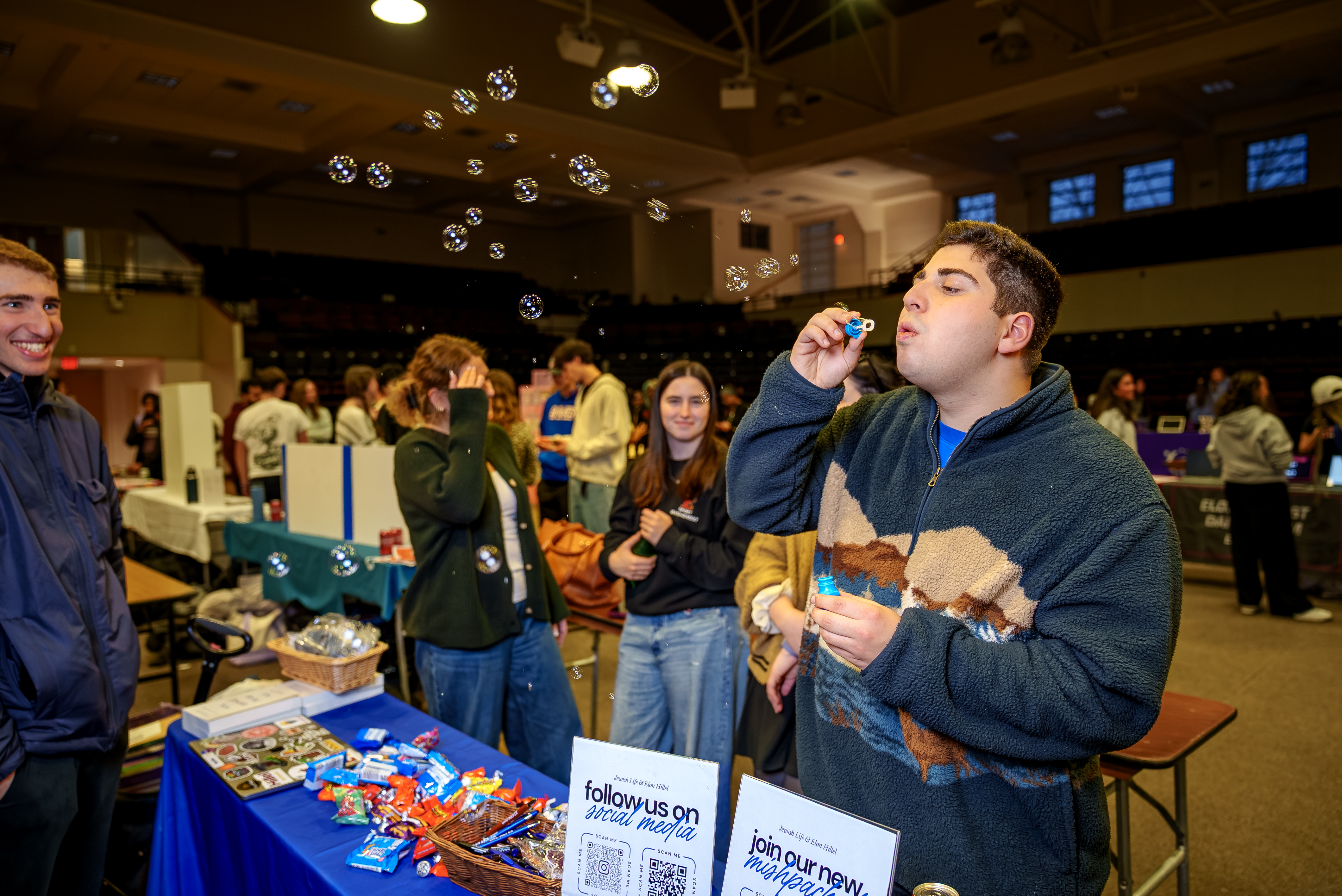 A student blows bubbles at an indoor campus event while others stand nearby at a table with candy and informational signs. The scene captures a lively, social atmosphere in a large hall with multiple activity tables in the background.
