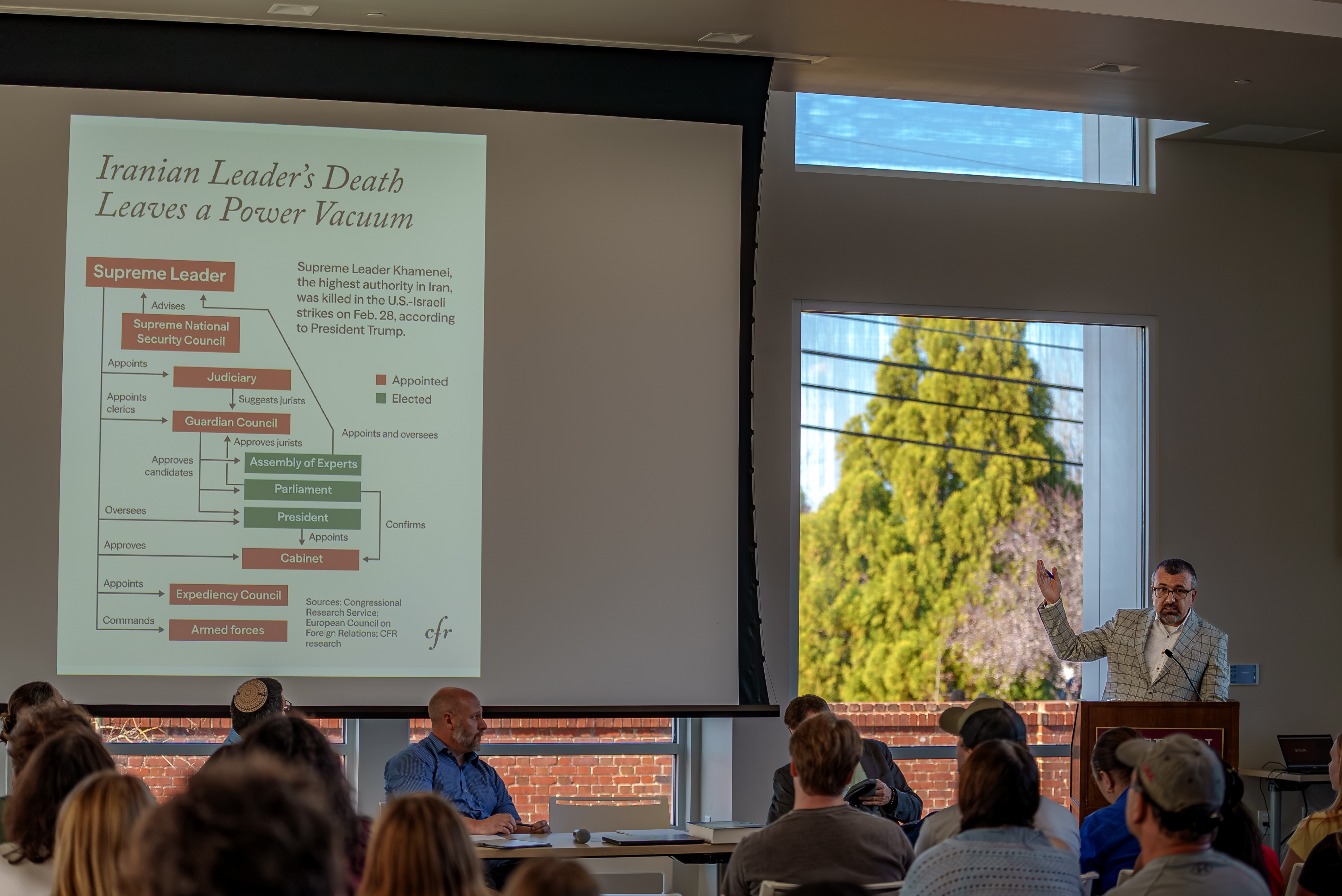 A presenter gestures toward a projected slide titled “Iranian Leader’s Death Leaves a Power Vacuum” while speaking to an audience seated in a lecture hall.