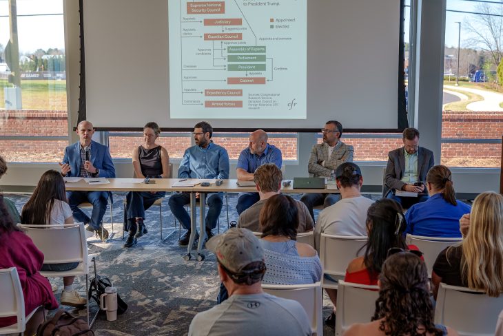 Several panelists sit at a long table addressing an audience while a slide about Iran’s political structure is projected on a screen behind them.