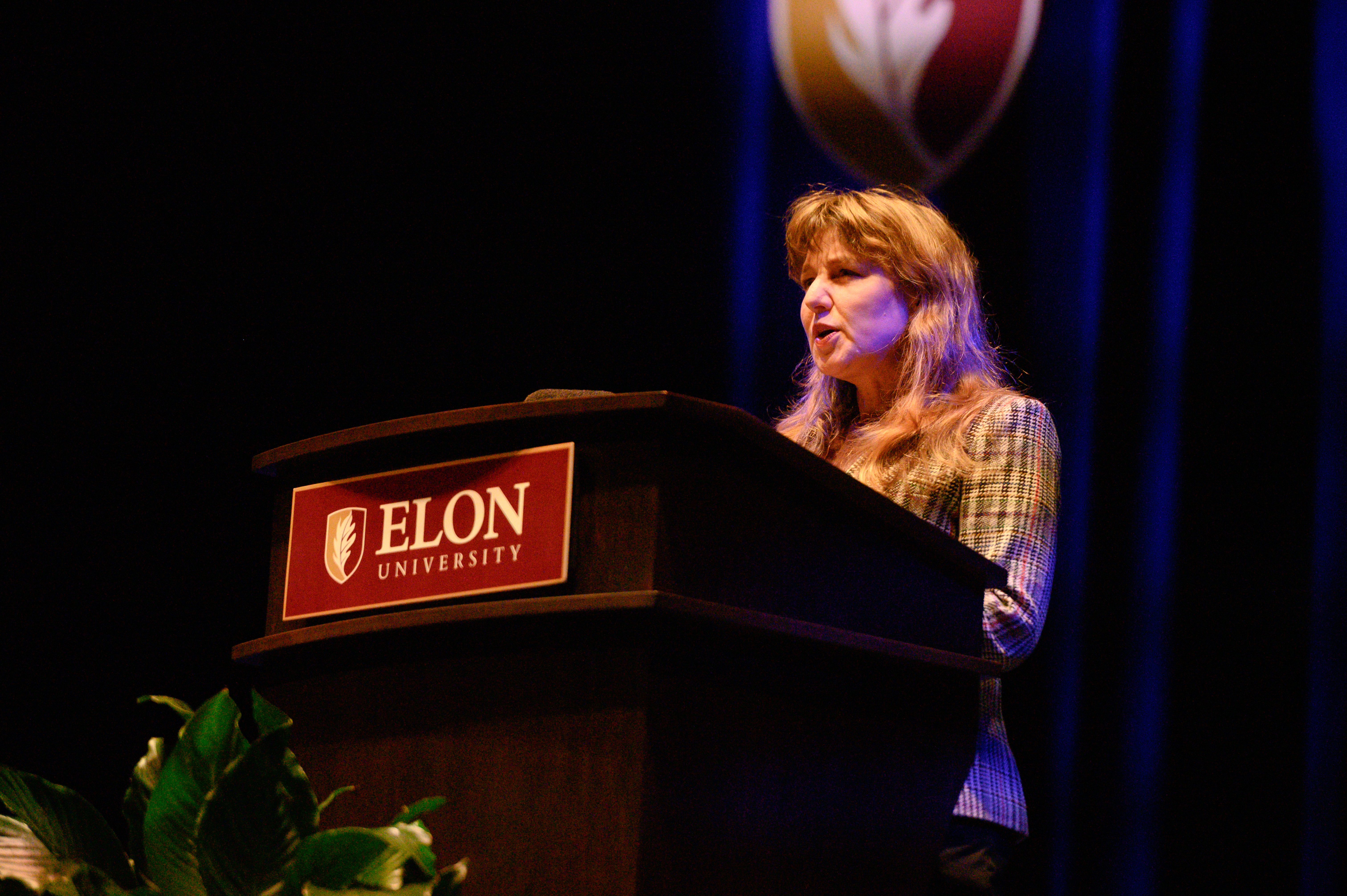 A speaker stands at the same podium delivering remarks during a formal event at Elon University.