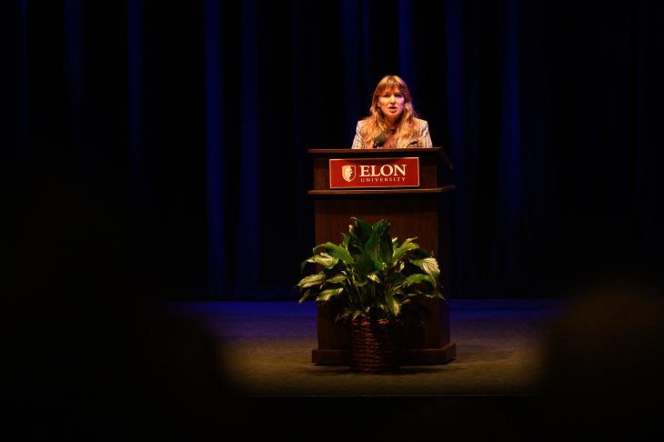 A wide view shows a speaker addressing an audience from the Elon University podium on a dimly lit stage with a plant in front.
