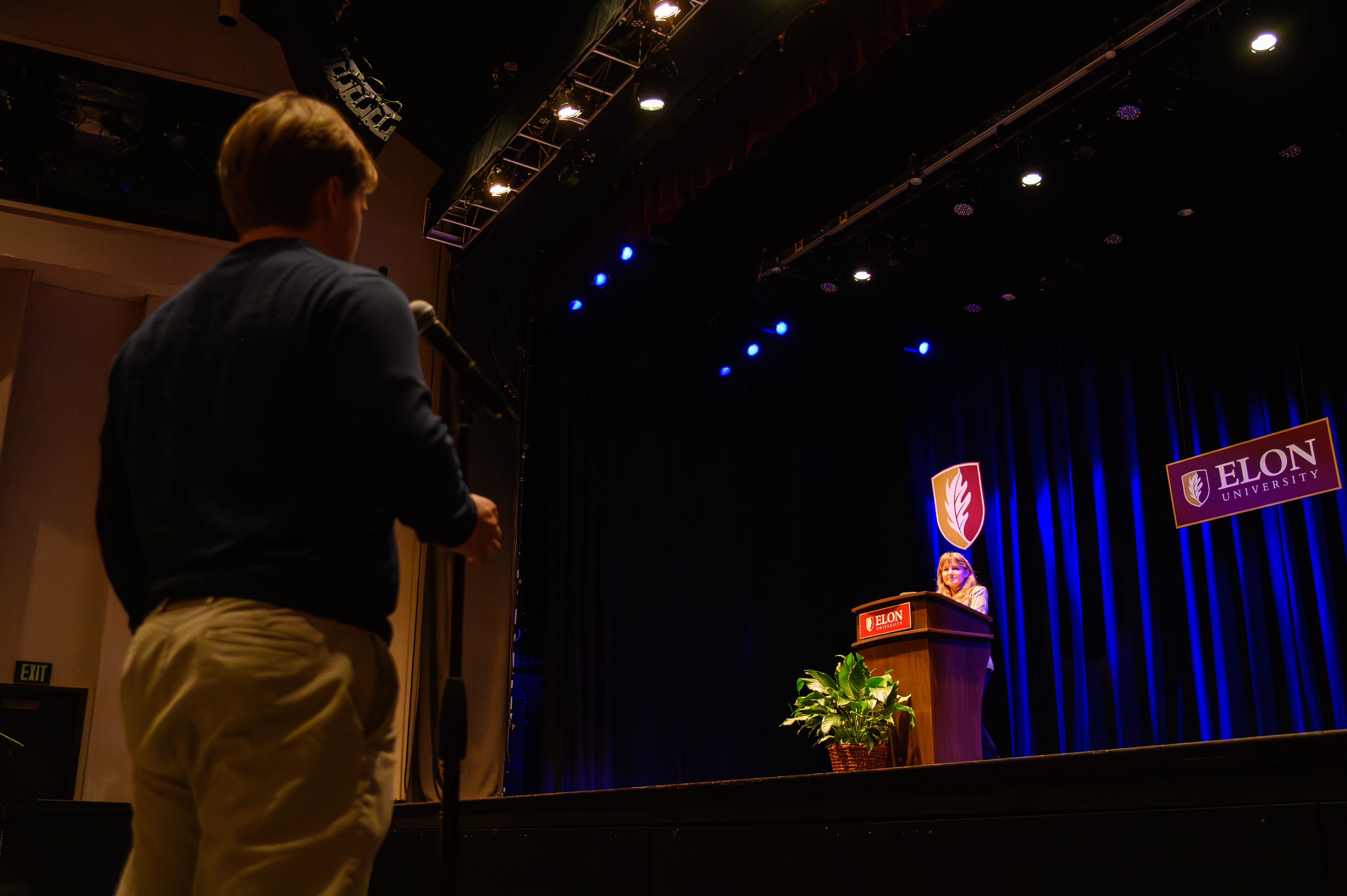A person stands at a microphone in the audience while a speaker addresses the room from an Elon University podium on stage. Blue stage lighting and university banners frame the formal event.