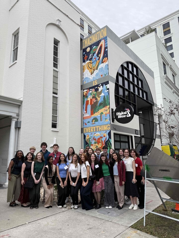 A large group of students pose together outside a building with a colorful mural reading “Imagination is Everything,” smiling for a group photo.