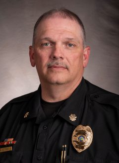 Doug has short brown hair with a thin mustache. He wears multiple lapels on his color and has a police badge on.