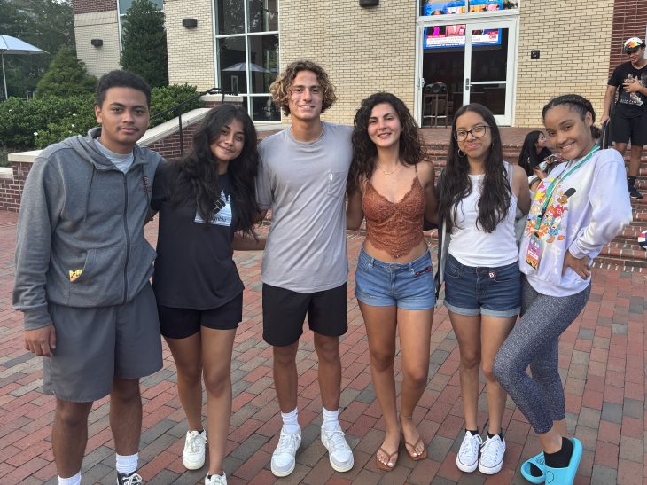 Six students stand together outdoors on a brick walkway, smiling with their arms around each other in a casual campus setting.