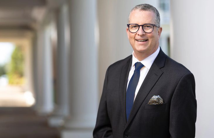 Jeff Stein has short hair and black rim glasses. He wears a black blazer and blue tie as he stands in front of pillars.