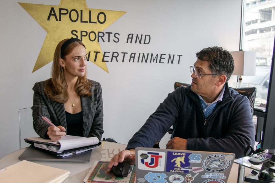 A man and woman seated behind a large desk facing each other. She is taking notes. Apollo Sports & Entertainment and a gold star painted on the wall behind them.
