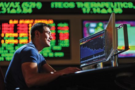 A man works at a computer displaying stock charts, with colorful market data boards glowing in the background.