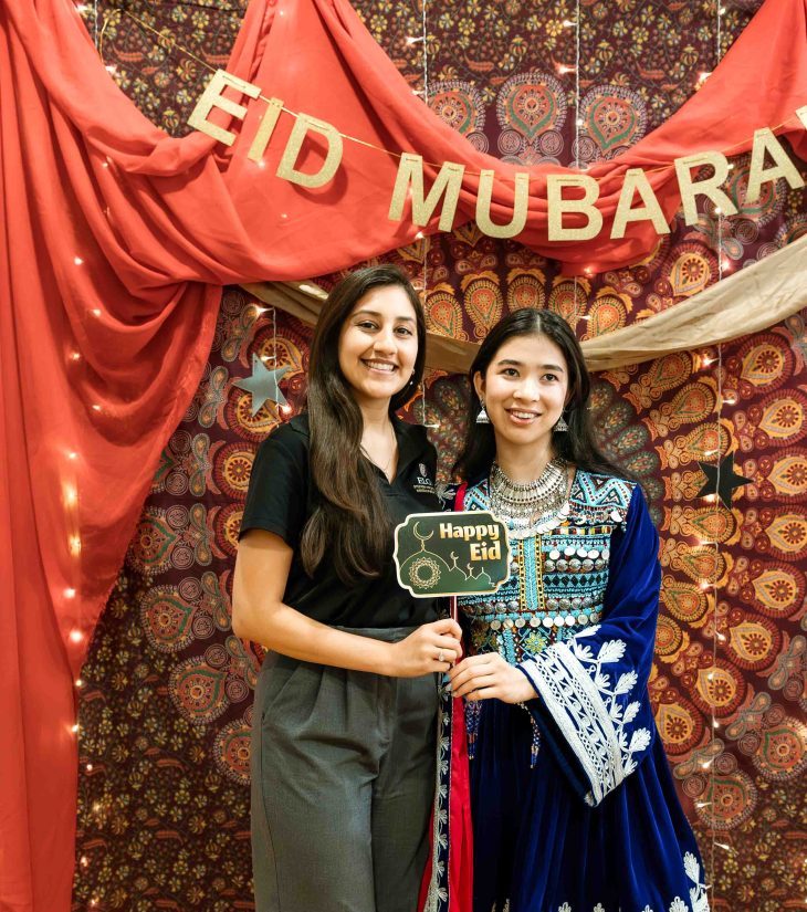 Two women smile in front of a colorful backdrop with an “Eid Mubarak” banner, one holding a “Happy Eid” sign.