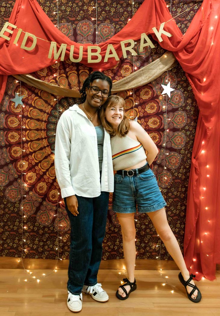 Two friends pose together in front of the same festive Eid backdrop decorated with lights and draped fabric.