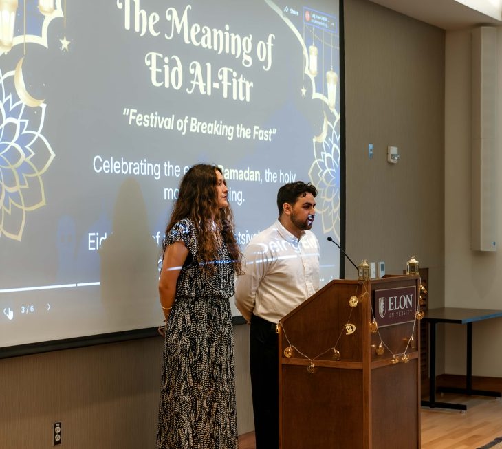 Two presenters stand at a podium in front of a screen explaining the meaning of Eid al-Fitr.
