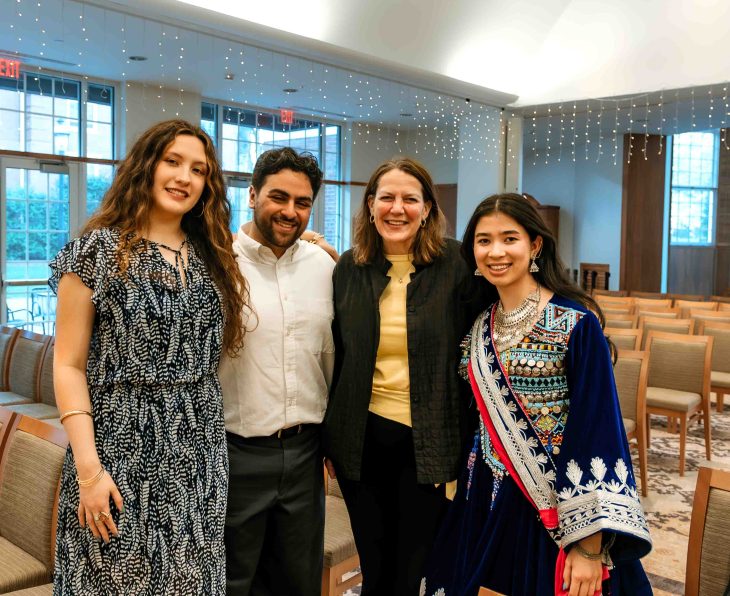 Four people stand together indoors, smiling in a well-lit room with chairs behind them.