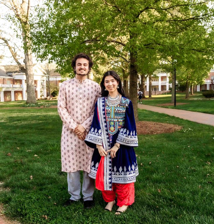 A man and woman in traditional clothing stand side by side on a grassy campus with trees and buildings in the background.