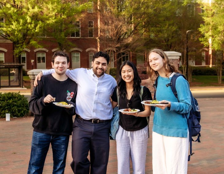 Four students pose together smiling on a college campus. Some holding plates of food.